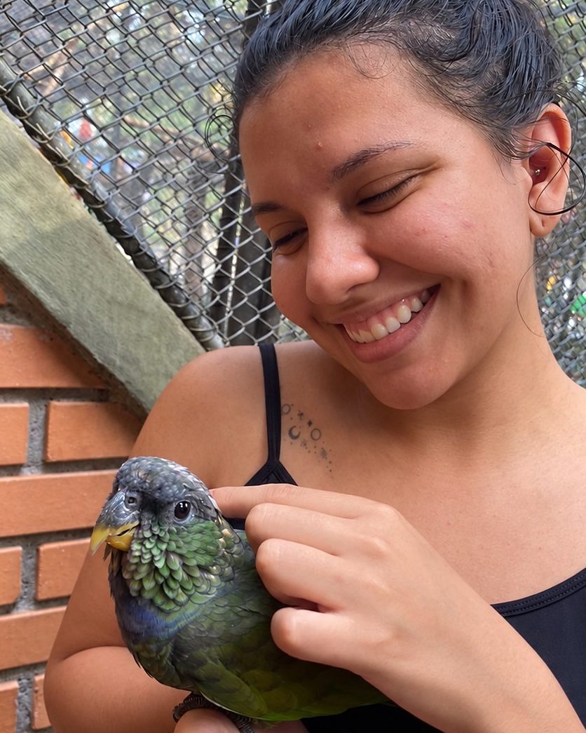 Bianca Barbara sorrindo com uma maitaca, ave verde, junto de um viveiro